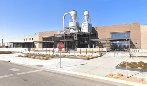 Exterior view of Sawmill Market, a modern building with large windows and two silos, set against a clear blue sky.