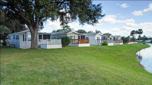 A row of mobile homes along a grassy area by a calm pond, with trees and a blue sky in the background.