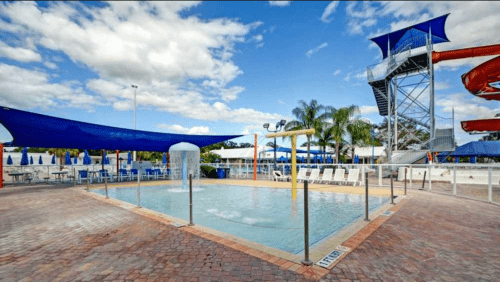 A sunny pool area with water slides, lounge chairs, and shaded seating under blue skies and scattered clouds.