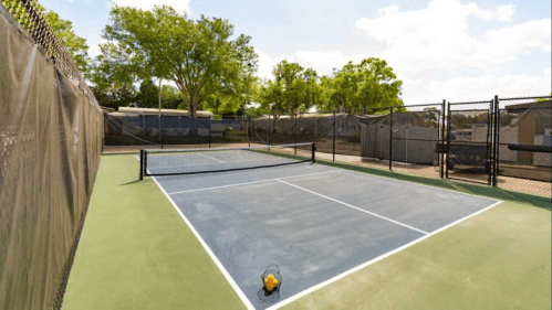 Two empty tennis courts surrounded by trees and a fence under a clear blue sky.