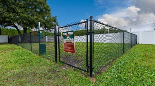 A fenced dog park with a sign, surrounded by green grass and trees under a blue sky.