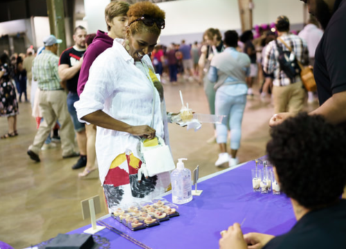 A woman in a white shirt smiles while holding a dessert at a busy event with people in the background.