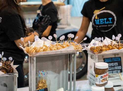 A display of chocolate-covered treats on sticks, with labels, at a food stall. Two people in black shirts are in the background.