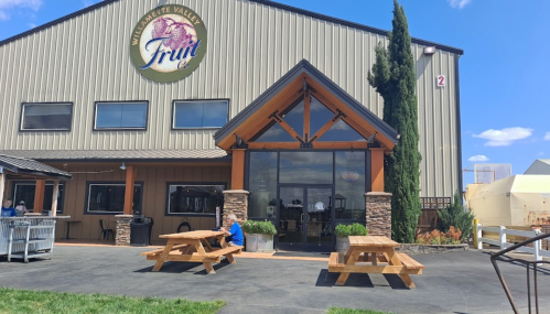 Exterior of a winery with a large sign, wooden entrance, and picnic tables in front on a sunny day.