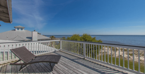 A sunny balcony with a lounge chair overlooking a calm beach and water under a clear blue sky.
