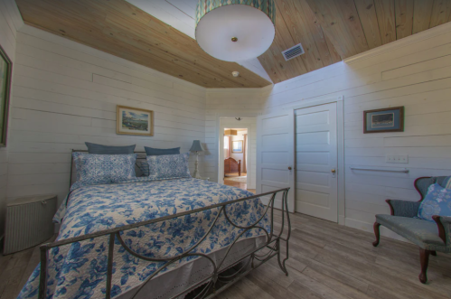 Cozy bedroom with a floral bedspread, wooden ceiling, and a vintage chair, featuring soft lighting and a warm atmosphere.