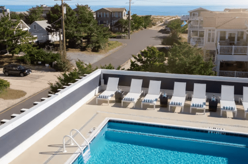 A rooftop pool with lounge chairs overlooks a beachside street and nearby houses.