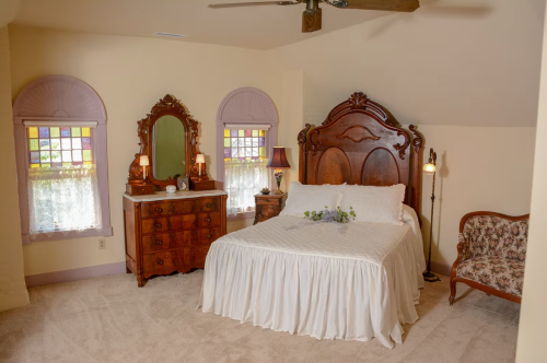 Cozy bedroom with a wooden bed, vintage dresser, and floral armchair, featuring stained glass windows and soft lighting.