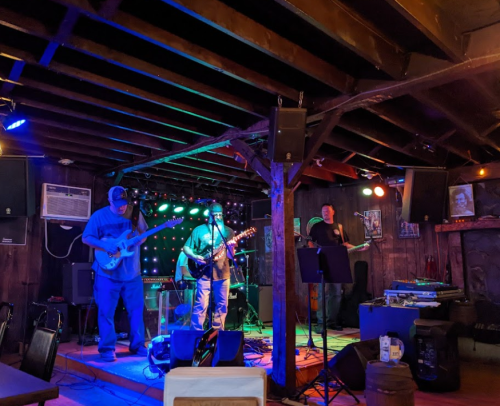 A band performs on stage in a dimly lit venue with colorful lights and wooden beams.