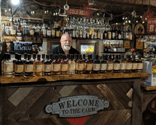 A man stands behind a wooden bar displaying various bottles of whiskey, with festive decorations in a rustic setting.