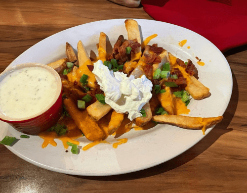 A plate of loaded fries topped with cheese, bacon, green onions, and sour cream, served with a side of ranch dressing.