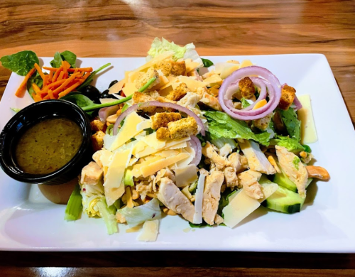 A colorful salad with grilled chicken, cheese, croutons, and assorted vegetables, served with a dressing on the side.