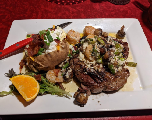 A plate featuring a grilled steak topped with shrimp and asparagus, alongside a baked potato with toppings and an orange slice.