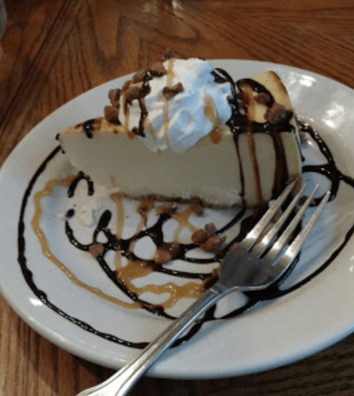 A slice of cheesecake topped with whipped cream, chocolate, and caramel drizzle on a plate with a fork.