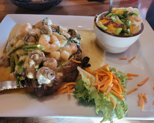 A plate featuring grilled steak topped with shrimp, mushrooms, and sauce, alongside a baked potato and salad.