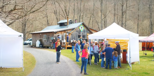 A lively outdoor event at a rustic venue, with people gathered around tents and a wooden building in a wooded area.