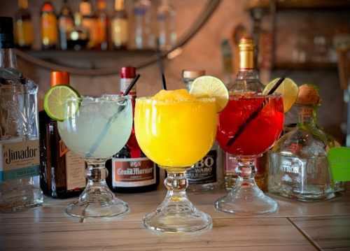 Three colorful cocktails in large glasses, garnished with lime slices, set against a backdrop of various liquor bottles.