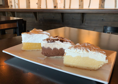 A plate with three slices of layered desserts: lemon, chocolate, and vanilla cream, on a black table.