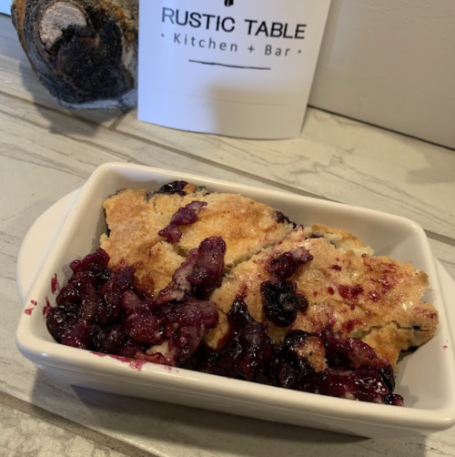 A white dish filled with berry cobbler, featuring a golden crust and vibrant berry filling, on a rustic table.