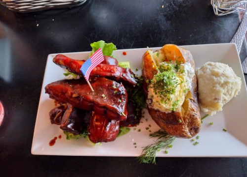 A plate featuring BBQ ribs, a baked potato with sour cream, and mashed potatoes, garnished with chives and a small American flag.