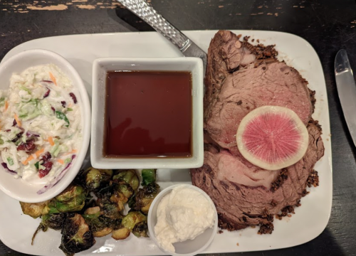 A plate with sliced roast beef, a side of coleslaw, Brussels sprouts, and a small bowl of sauce.