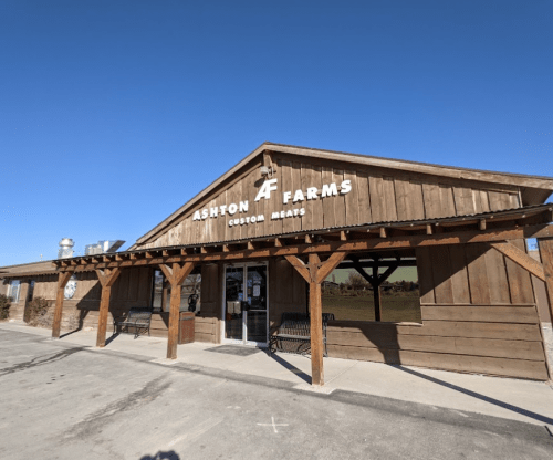 Exterior of Ashton Farms, a rustic wooden building with a sign for custom meats under a clear blue sky.