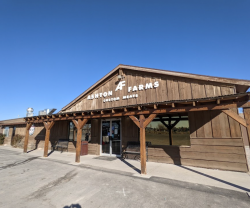 Exterior of Ashton Farms, a rustic wooden building with a sign for custom meats under a clear blue sky.
