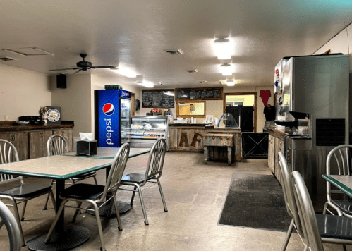 A cozy café interior with metal tables, a Pepsi fridge, and a display case, featuring rustic decor and a welcoming atmosphere.