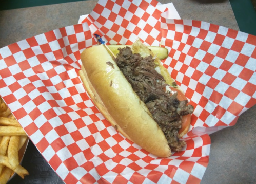 A sandwich filled with shredded beef on a hoagie roll, served with pickles, on a checkered paper plate. French fries in the background.