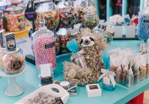 A colorful candy shop display featuring jars of sweets, a plush sloth, and assorted treats on a bright blue table.