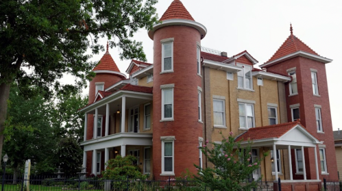 A large, historic house with red brick and turreted towers, featuring a wraparound porch and lush greenery.