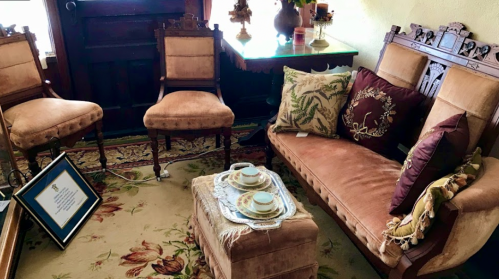 A cozy sitting area with vintage furniture, decorative pillows, a small table, and a framed certificate on the floor.