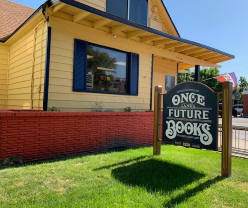 A yellow house with blue shutters and a sign reading "Once and Future Books" in front, surrounded by green grass.