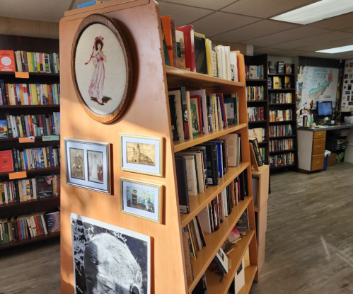 A wooden bookshelf filled with books, featuring framed photos and a decorative embroidery piece in a cozy bookstore.