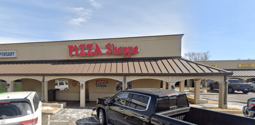 A storefront with a red sign reading "PIZZA Shoppe" under a metal roof, surrounded by parked vehicles.