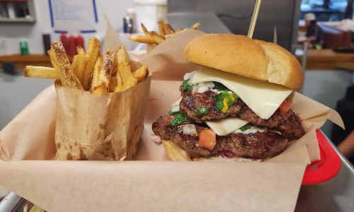 A stacked burger with cheese, onions, and tomatoes, served with a side of crispy fries in a paper container.