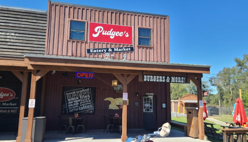 Exterior of Pudgee's Eatery & Market, featuring a wooden facade, "Open" sign, and outdoor seating with umbrellas.