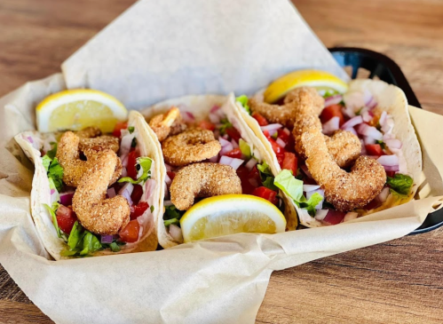 Three crispy shrimp tacos with lettuce, tomatoes, and onions, garnished with lemon wedges, served in a basket.