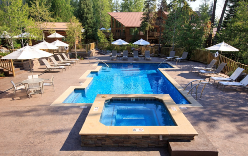A clear blue pool with a hot tub, surrounded by lounge chairs and umbrellas, set in a wooded area.