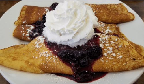 Three crepes topped with whipped cream and berry sauce, dusted with powdered sugar, served on a white plate.