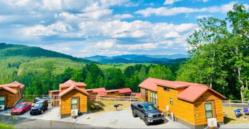 A scenic view of wooden cabins surrounded by lush green mountains and a blue sky with fluffy clouds.