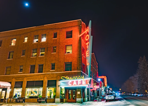 A brightly lit vintage hotel and café at night, with neon signs glowing against a dark sky.