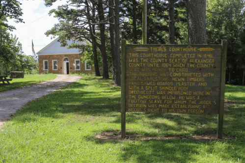 Historic courthouse from 1848 with a sign detailing its history, surrounded by trees and a gravel path.