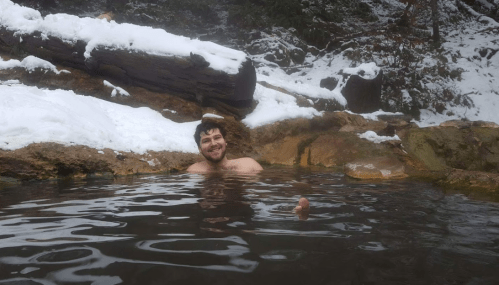 A person smiles while relaxing in a natural hot spring surrounded by snow-covered rocks and trees.