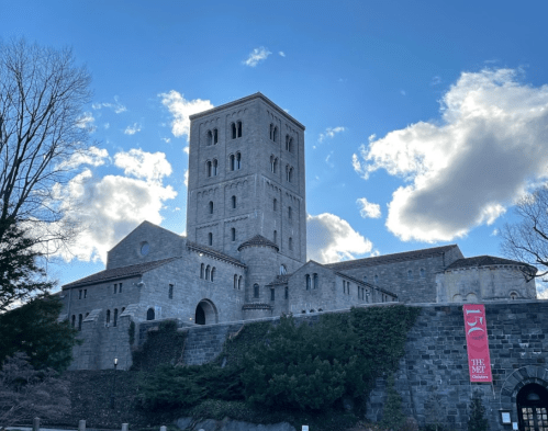 A tall stone castle-like building under a blue sky with clouds, surrounded by trees and greenery.