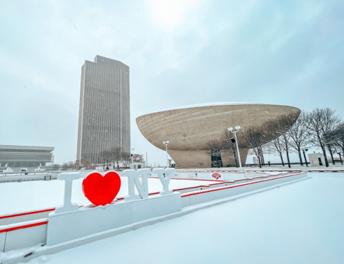 Snow-covered scene featuring a large "I ❤️ NY" sign, modern architecture, and a cloudy sky.