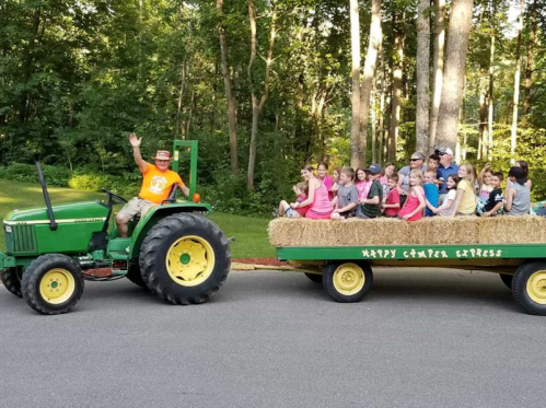 A man waves from a green tractor pulling a hayride filled with children through a wooded area.