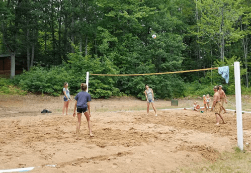 A group of people playing beach volleyball on a sandy court surrounded by trees.