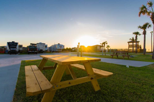 A wooden picnic table in a grassy area with RVs and a sunset in the background.
