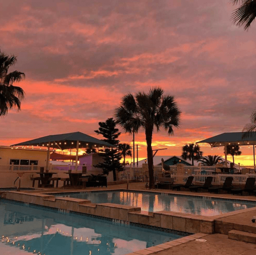 A serene poolside scene at sunset, with vibrant orange and pink skies and palm trees silhouetted against the horizon.